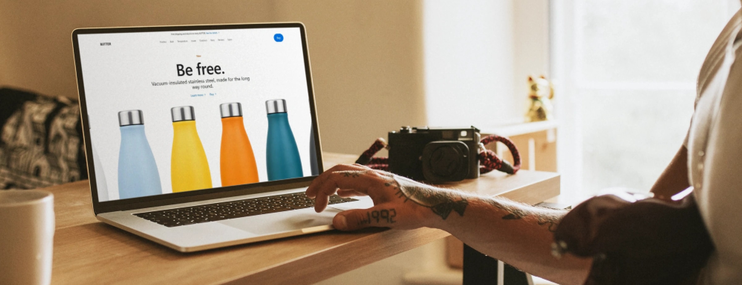 A hand reaching for the trackpad of a laptop showing the B3TTER site, camera and coffee cup nearby